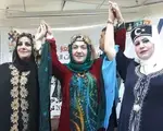 Photo of three Iraqi women from different ethnic backgrounds in traditional dress raising their hands together in solidarity.