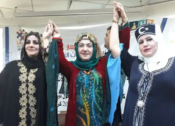 Photo of three Iraqi women from different ethnic backgrounds in traditional dress raising their hands together in solidarity.